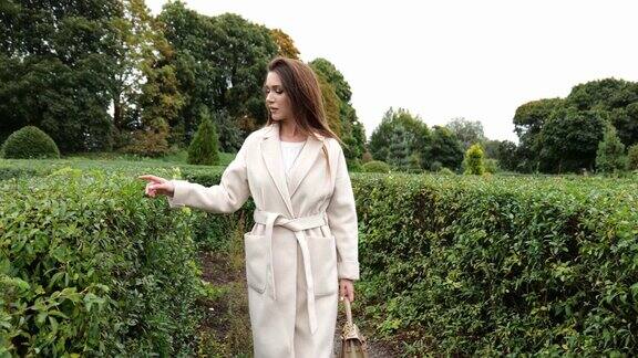 一位身穿米色外套的褐发年轻女子走在植物园的绿色灌木丛旁一个美丽女人的肖像
