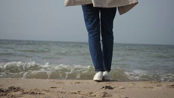 一个年轻的女人在米色外套和蓝色牛仔裤跳舞的海滩上的特写沿着海岸走