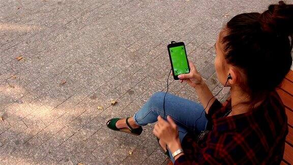 年轻漂亮的女孩坐在公园的长椅上用智能手机浏览社交网络