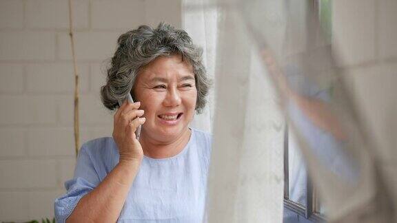 肖像高级亚洲妇女在家里的窗户边用智能手机愉快地交谈和微笑