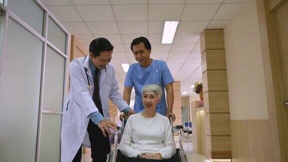 亚洲资深男医生和助理在医院走廊上推着轮椅与老年妇女病人行走交谈