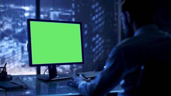 深夜一个商人在他的私人办公室里用一台带有绿色模拟屏幕的个人电脑工作可以看到大城市的窗户