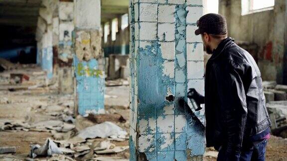 一个留着胡子的涂鸦艺术家正在一幢废弃建筑的柱子上用明亮的喷雾剂喷漆空无一人的工业建筑肮脏的墙壁和破碎的窗户