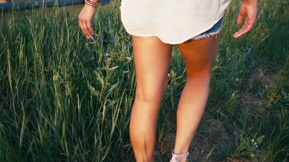 年轻美丽的女人在运动鞋和短裤走在草地与绿色的草自然慢动作视频女孩在田野的腿在日落近景在草地上的阳光生活剪影