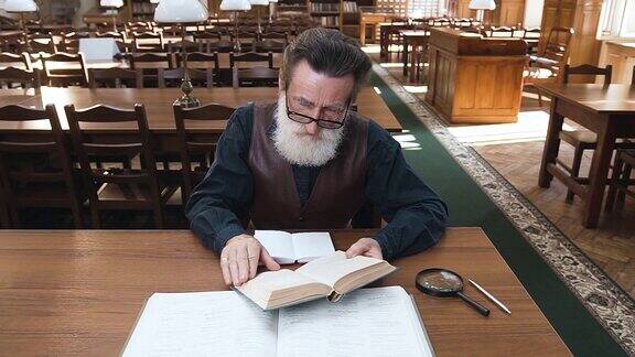 有魅力专注读书的大胡子老人戴着眼镜坐在图书馆的桌子上拿着书