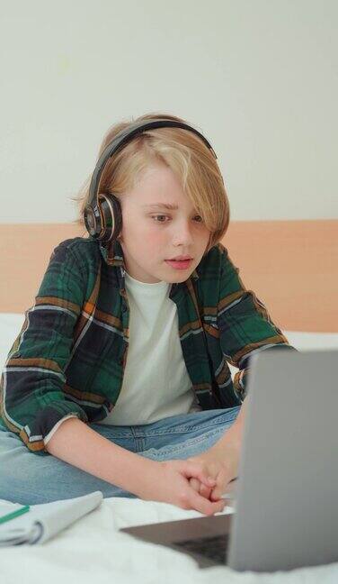 十几岁的男孩使用笔记本电脑和耳机孩子在床上看在线视频课程聊天视频会议男生学习做作业电子学习远程教育课程垂直