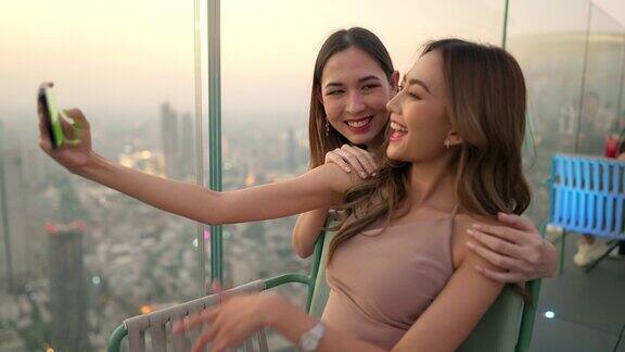 日落时分4K的亚洲女性朋友在摩天大楼的屋顶餐厅一起用智能手机自拍