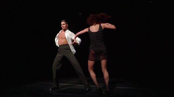 两名交谊舞舞者从黑暗中走出来来到泛光灯光下的舞台上他们迈着灵活的步伐挥舞着手臂按照拉丁美洲舞蹈的风格旋转着臀部男人让女人翻筋斗女人坐下来做深弓步