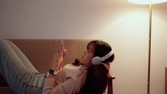 美丽的年轻亚洲女人拿着她的智能手机正在沙发上播放音乐