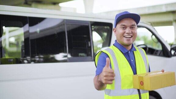 快邮配送服务理念年轻的亚洲快递员戴着蓝帽子站在一辆白色汽车前一只手拿着包裹拇指对着镜头4k决议