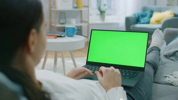 一个女人躺在家里的沙发上用绿色模拟屏幕的笔记本电脑工作用电脑上网和浏览社交网络的女孩过肩镜头拍摄