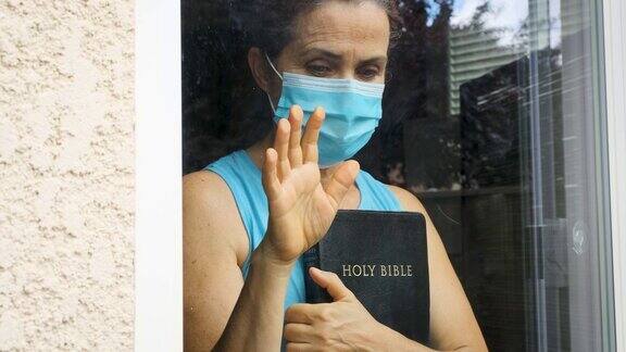 一个成熟的女人拿着一本圣经戴着外科口罩从窗户往外看
