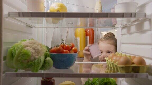 可爱的小女孩打开冰箱门从里面拿出健康的酸奶聪明的小孩吃得健康厨房冰箱里的摄像头从装满食物的冰箱拍摄的观点