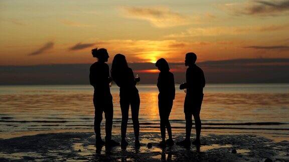 后视图剪影四个不同的年轻朋友站在海边的海滩上看着美丽的橙色日落喝啤酒从玻璃瓶和聊