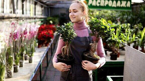 年轻的女花匠扎着马尾辫在花店或花房的花丛中走着手里拿着两个花盆微笑着看着镜头Slowmotion拍摄