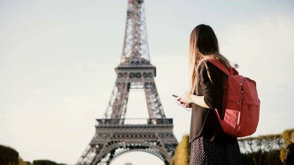 年轻漂亮的女子背着双肩包站在埃菲尔铁塔附近用智能手机上网