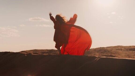 一位身穿红色连衣裙的亚洲妇女在日落时的沙丘上迎着太阳跳舞