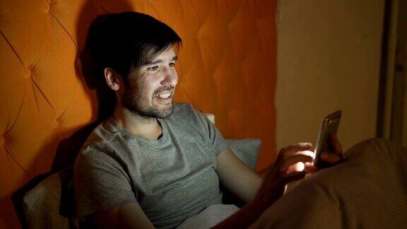 年轻微笑的男子晚上躺在家里的床上用智能手机浏览社交媒体