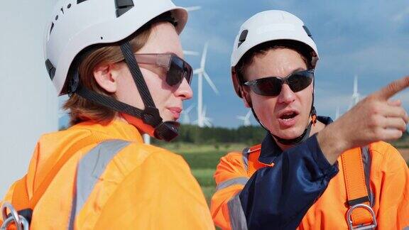 两名身穿安全服的电气工程师在风力发电机场边指着一台风力发电机进行合作讨论用数字平板检查涡轮系统专业人士利用风力发电来生产清洁电力