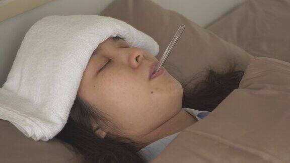 侧视图摇摄生病的亚洲妇女与毛巾在额头上以减轻发烧和降低体温躺在床上睡觉口腔温度计在她的嘴里