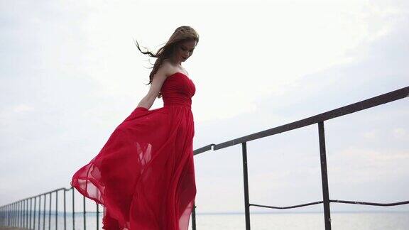 在刮风的天气里一个穿着红色长裙的漂亮女人站在海边的码头上