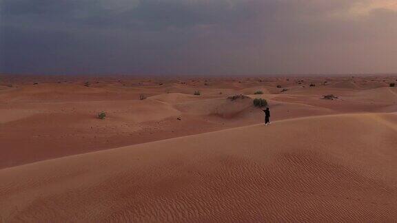 一架无人机在一名身穿阿拉伯联合酋长国传统服装的女子旁边的空中俯瞰她正在沙漠中的沙丘上行走