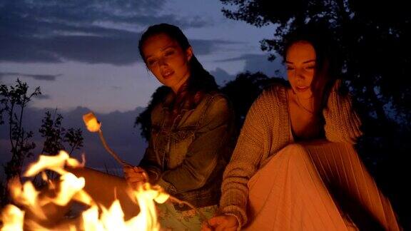 两个年轻的白人女孩晚上坐在篝火旁准备一个棉花糖看着篝火思考着