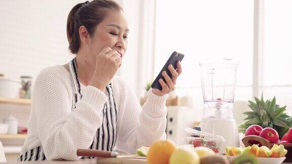 亚洲资深女性一边吃水果一边用手机感到幸福准备用不同的口味和酸奶自制水果冰沙健康天然益生菌调味饮料在厨房学习使用科技