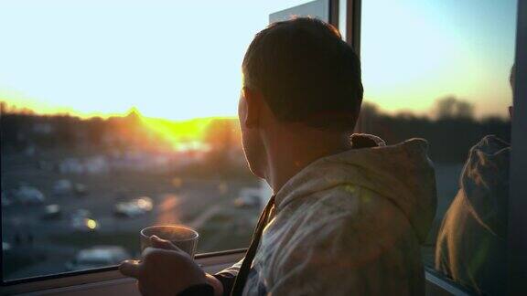 在美丽的日落时分年轻人站在开着的窗边端着一杯热茶欣赏着风景