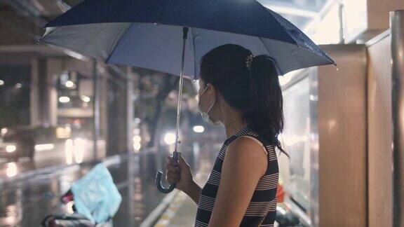 一名亚洲女子撑着伞在公交车站等车