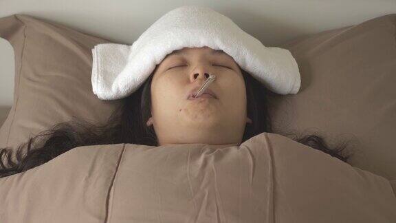 正面图将患病的亚洲妇女用毛巾敷在额头以退烧降体温躺在床上睡觉嘴里含着体温计
