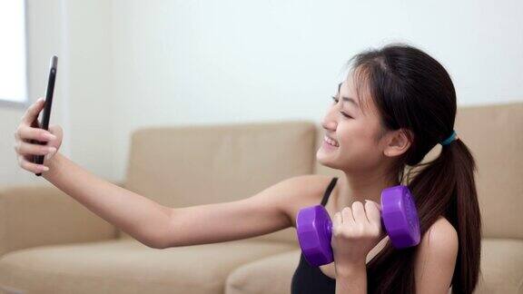 有吸引力的亚洲年轻健身女性在家里举哑铃举重锻炼使用智能手机直播在家里分享在社交媒体上女性训练和锻炼用手机拍照