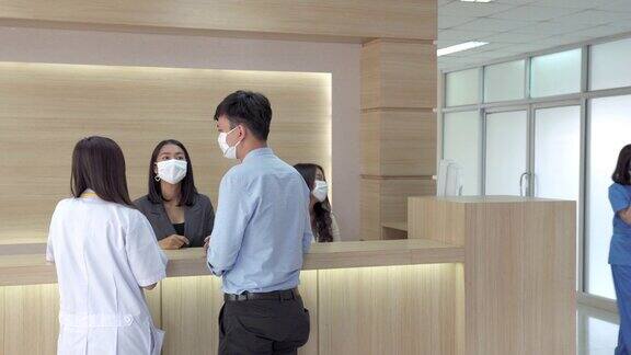 4K年轻亚洲男性患者佩戴医用口罩在现代医院保健设施大厅向女性接待和医生询问治疗信息医生和医疗保健服务理念