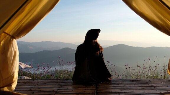 后视图的一个女人坐在木制阳台上而看日出和一个美丽的山景外的帐篷