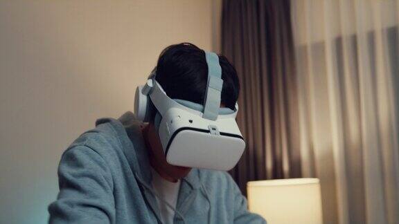 年轻的亚洲男性玩家穿着灰色的卫衣带着VR的oculus握着VR操纵杆控制器在晚上的客厅家里感受刺激和有趣的游戏新体验