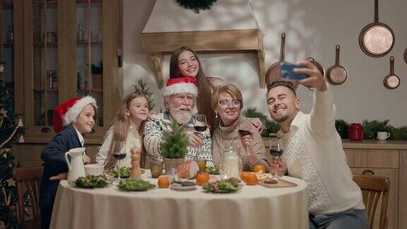 家庭年夜饭餐桌上一个戴圣诞帽的老人和一个戴眼镜的女人两个孩子一个男孩一个女孩两个年轻人用手机自拍的男人男人和女人拿着酒杯
