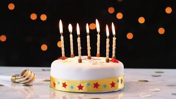 7支点燃的蜡烛放在一个装饰好的白色生日蛋糕上旁边有一个派对吹风机背景是散焦灯