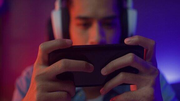 快乐亚洲男人游戏玩家戴耳机比赛玩在线电子游戏与智能手机彩色霓虹灯在客厅的夜晚现代房子