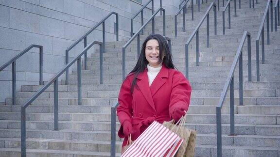 一幅欢快的白人女孩穿着红色外套在户外拿着购物袋跳舞的肖像兴奋的女性购物狂满意的购买快乐生活方式shopaholism