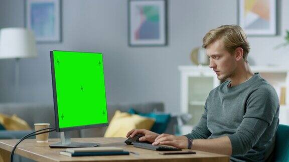 英俊专注的年轻人在一个绿色模拟屏幕个人电脑上工作而坐在他的办公桌在舒适的公寓