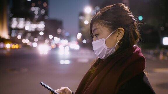 女商人晚上在城市等待路灯时使用智能手机并戴上防护口罩