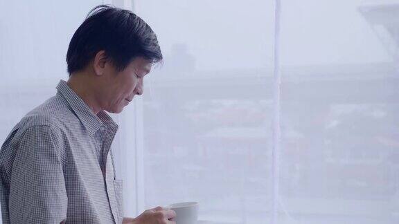 一位上了年纪的亚洲人站在窗前手里拿着咖啡杯面带平静的表情望着窗外