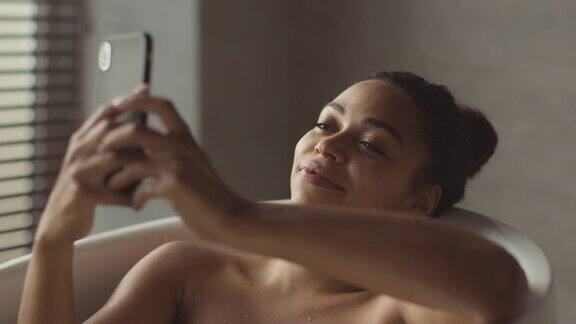 年轻可爱的非洲裔美国女士放松在浴室在家里在智能手机上上网社交媒体跟踪拍摄