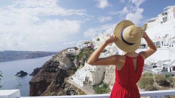 身穿红色连衣裙的妇女在圣托里尼岛享受假日旅行女孩游客手持太阳帽看着著名城市伊亚她正在参观一个著名的旅游景点红色的慢动作