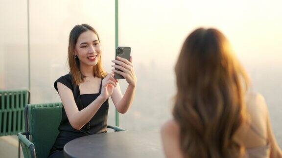 日落时分4K亚洲女性朋友在屋顶餐厅用智能手机拍照