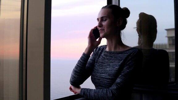 夕阳西下的海边一个微笑着的年轻女子拨通了电话号码开始打电话她站在窗前面带微笑背景是美丽的天空和大海