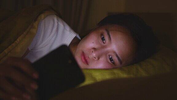 年轻的亚洲女性晚上在卧室的床上用智能手机上网和社交媒体