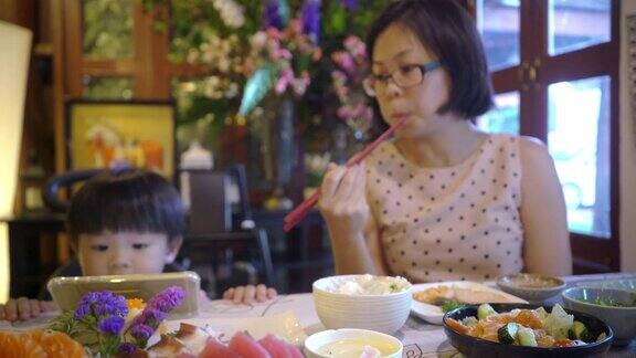 在一家日本餐厅里一位亚洲母亲正在挑选一块三文鱼生鱼片吃而她的儿子正在用智能手机玩游戏