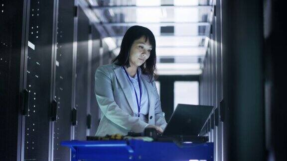 亚洲女IT工程师在一个大型数据中心满是机架服务器的应急手推车笔记本上工作