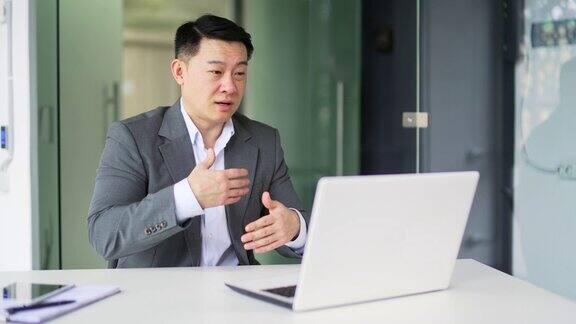 一位亚洲商人坐在现代办公室的办公桌前用笔记本电脑进行视频通话